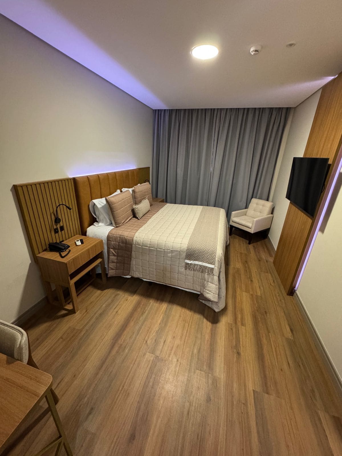 Quarto de hotel moderno com uma cama grande, cabeceira de madeira, mesa de cabeceira com abajur, uma cadeira e televisão na parede.