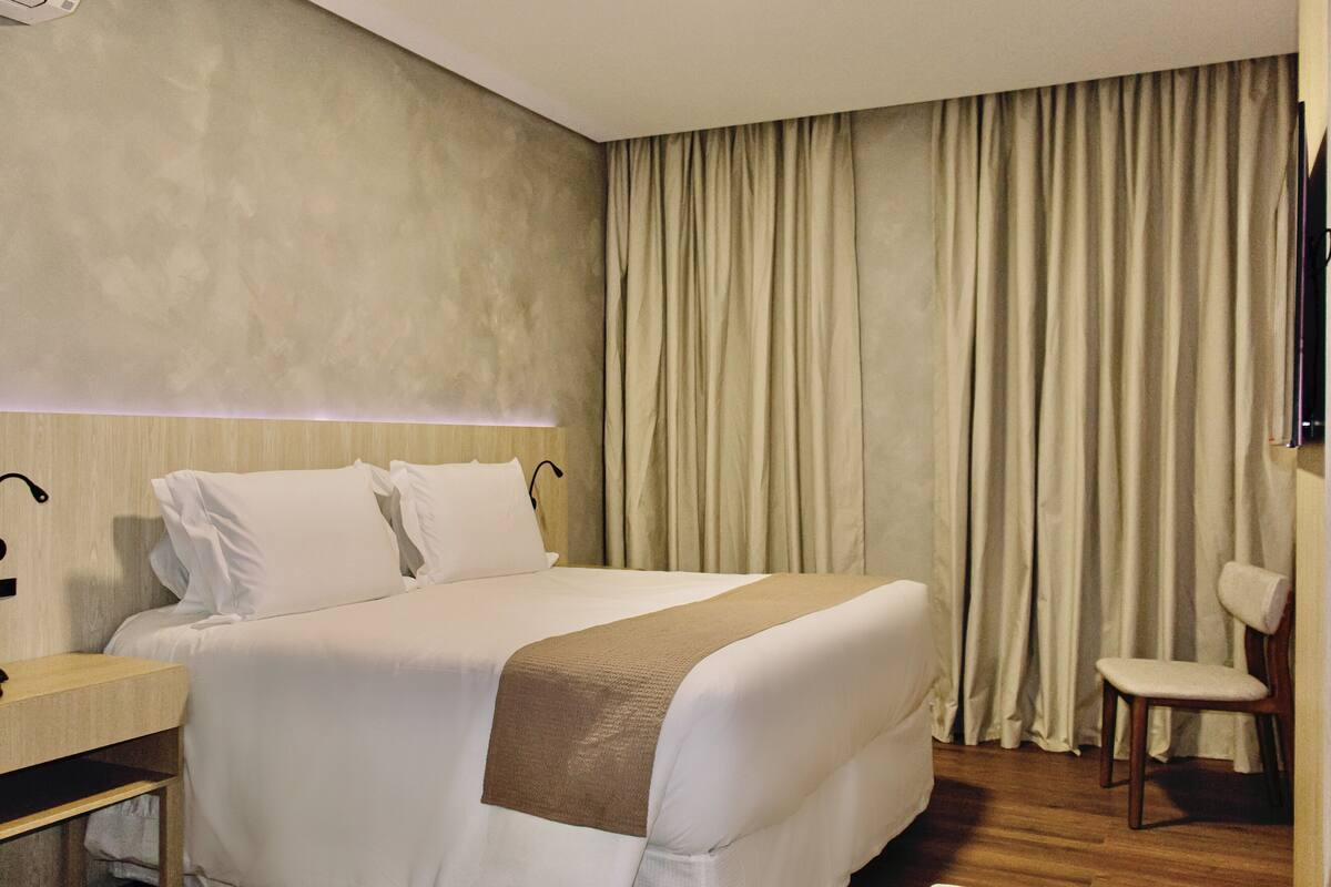 Een hotelkamer met een bed, een nachtkastje van hout, een stoel en een groot raam met gordijnen.