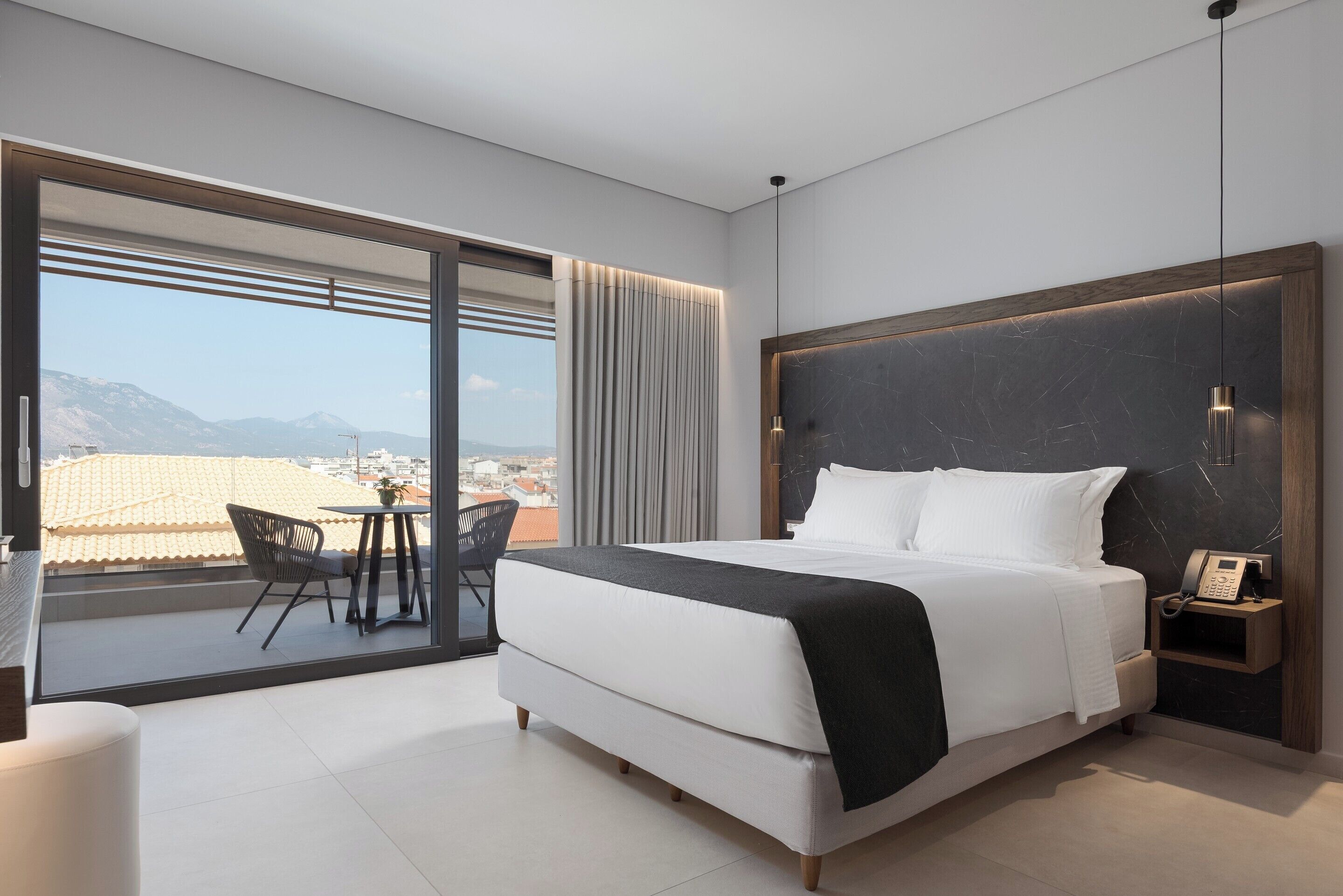 Habitación de hotel moderna con una cama grande, teléfono junto a la cama y vistas de la ciudad a través de ventanales de gran altura.