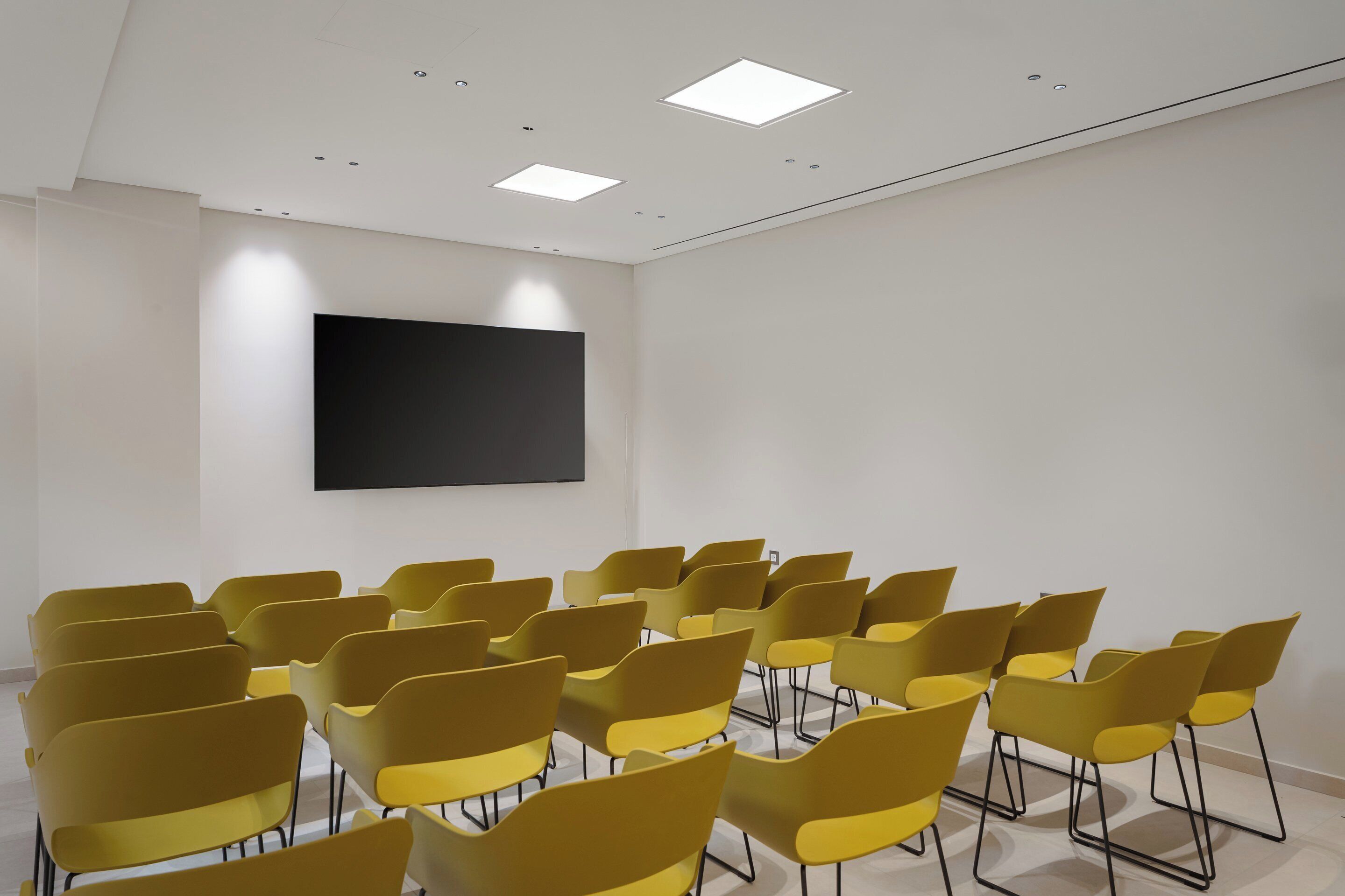 Un salón de conferencias con hileras de sillas amarillas, un televisor de pantalla plana grande en la pared y luces empotradas en el techo.