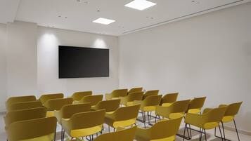 Un salón de conferencias con hileras de sillas amarillas, un televisor de pantalla plana grande en la pared y luces empotradas en el techo.