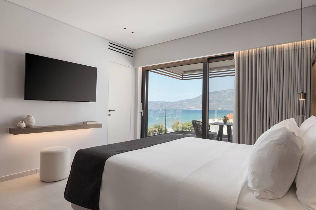 Ein modernes Hotelzimmer mit einem großen Bett, einem flachbildfernseher an der Wand, einem kleinen Tisch mit einem Stuhl und Meerblick durch große Fenster.