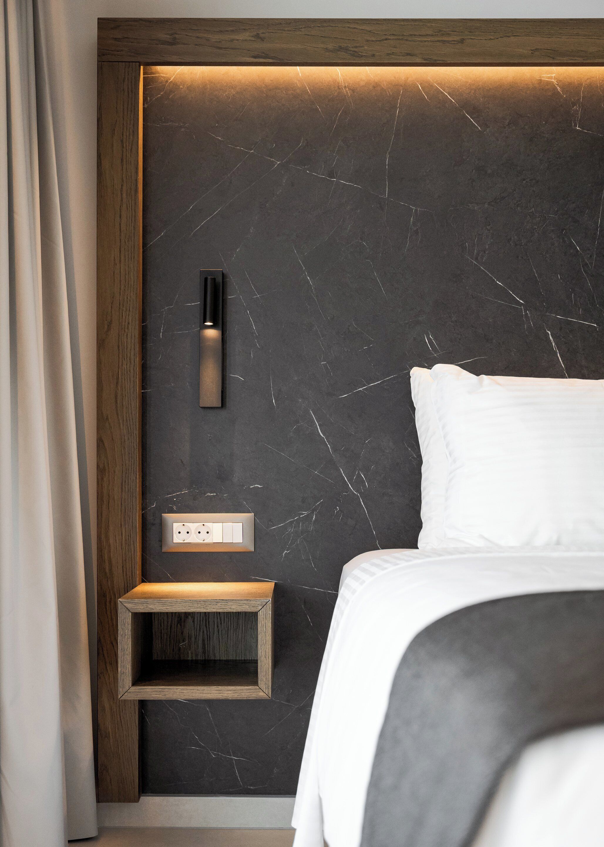 A modern bedroom with a dark marble wall, a bedside table, a wall-mounted light, and a bed with white bedding.