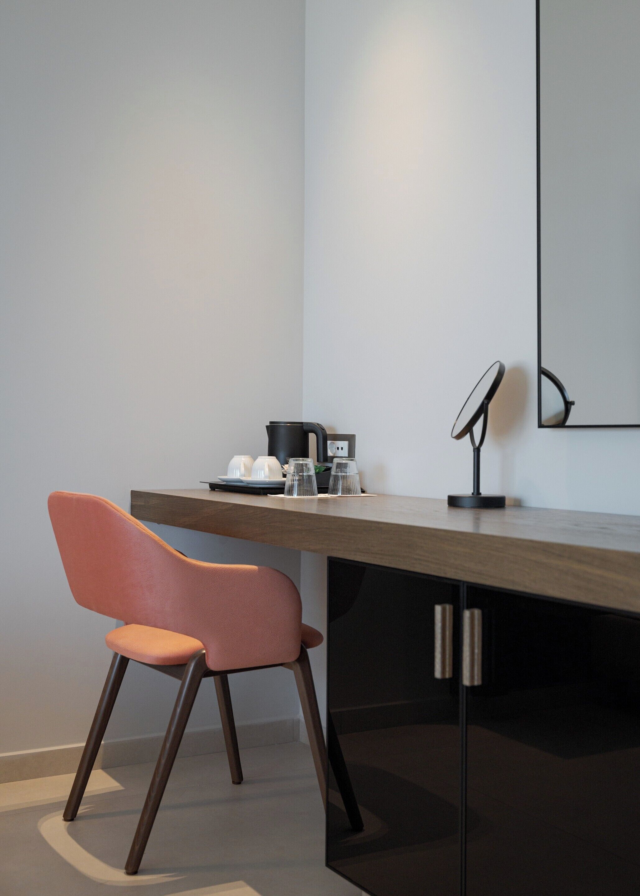 Een moderne kamer met een houten bureau, een roze stoel, een zwarte lamp en een spiegel aan de muur.