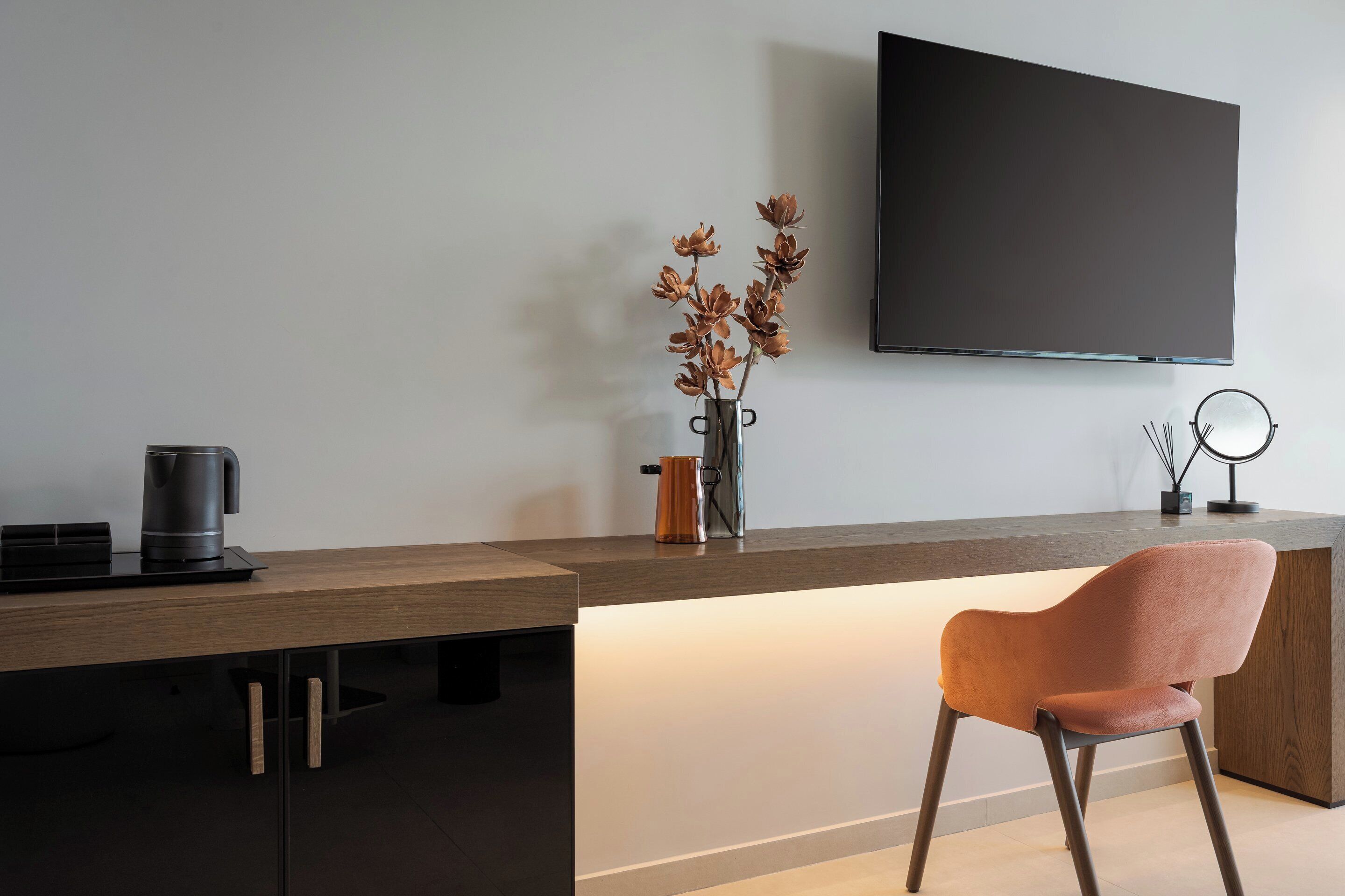 A modern kitchen with a wooden countertop, a coffee maker, a vase with dried flowers, and a flat-screen TV mounted on the wall.