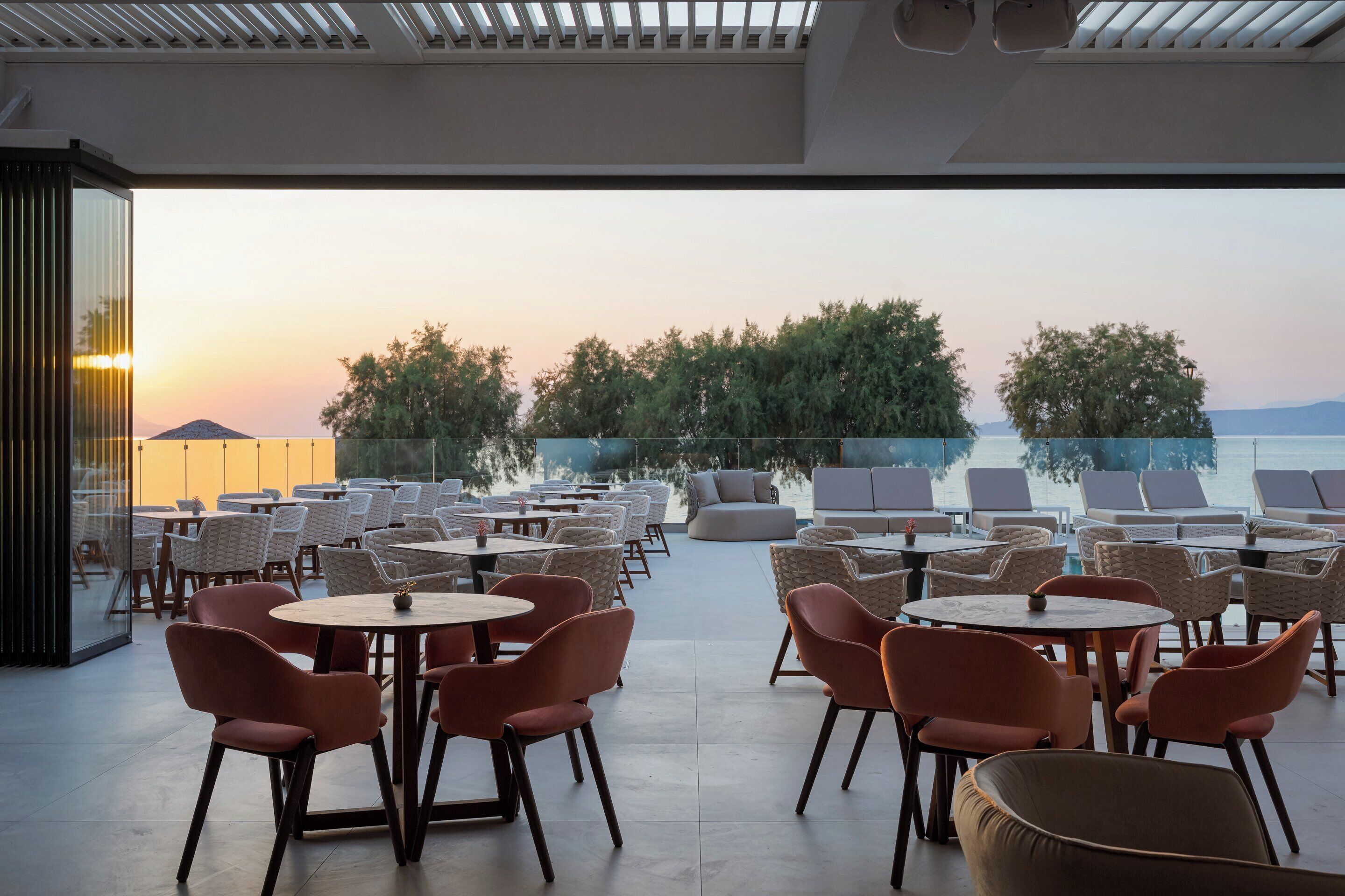 An outdoor seating area with round tables and chairs, overlooking a body of water and trees.