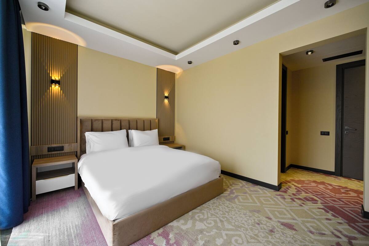 Una habitación de hotel con una cama grande, una mesita de noche, una lámpara de pared y una alfombra estampada.