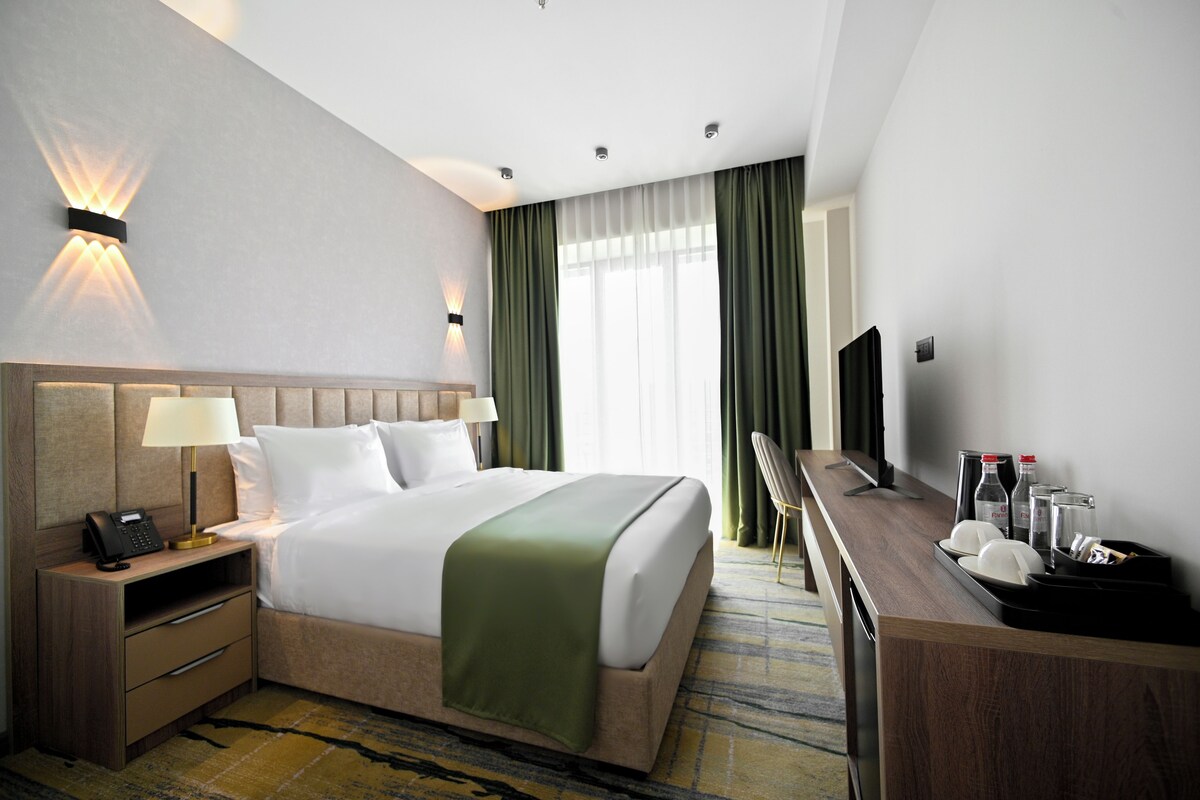 Quarto de hotel com cama, mesa de cabeceira com abajur, televisão e escrivaninha com um conjunto de objetos.