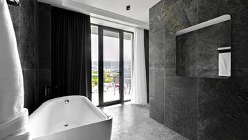 Un baño moderno con una bañera independiente, paredes de mármol oscuro y un amplio ventanal corredero que da a un balcón con vistas a la ciudad.