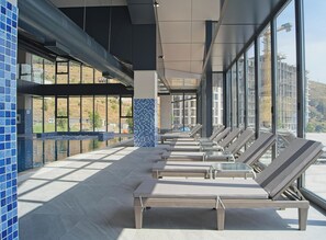 Un área de piscina interior moderna con sillones de descanso y vistas a una obra en construcción a través de amplios ventanales.