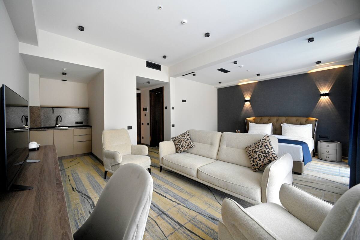 Una habitación de hotel moderna con sofá, sillones, televisor, zona de cocina y cama con cabecero.