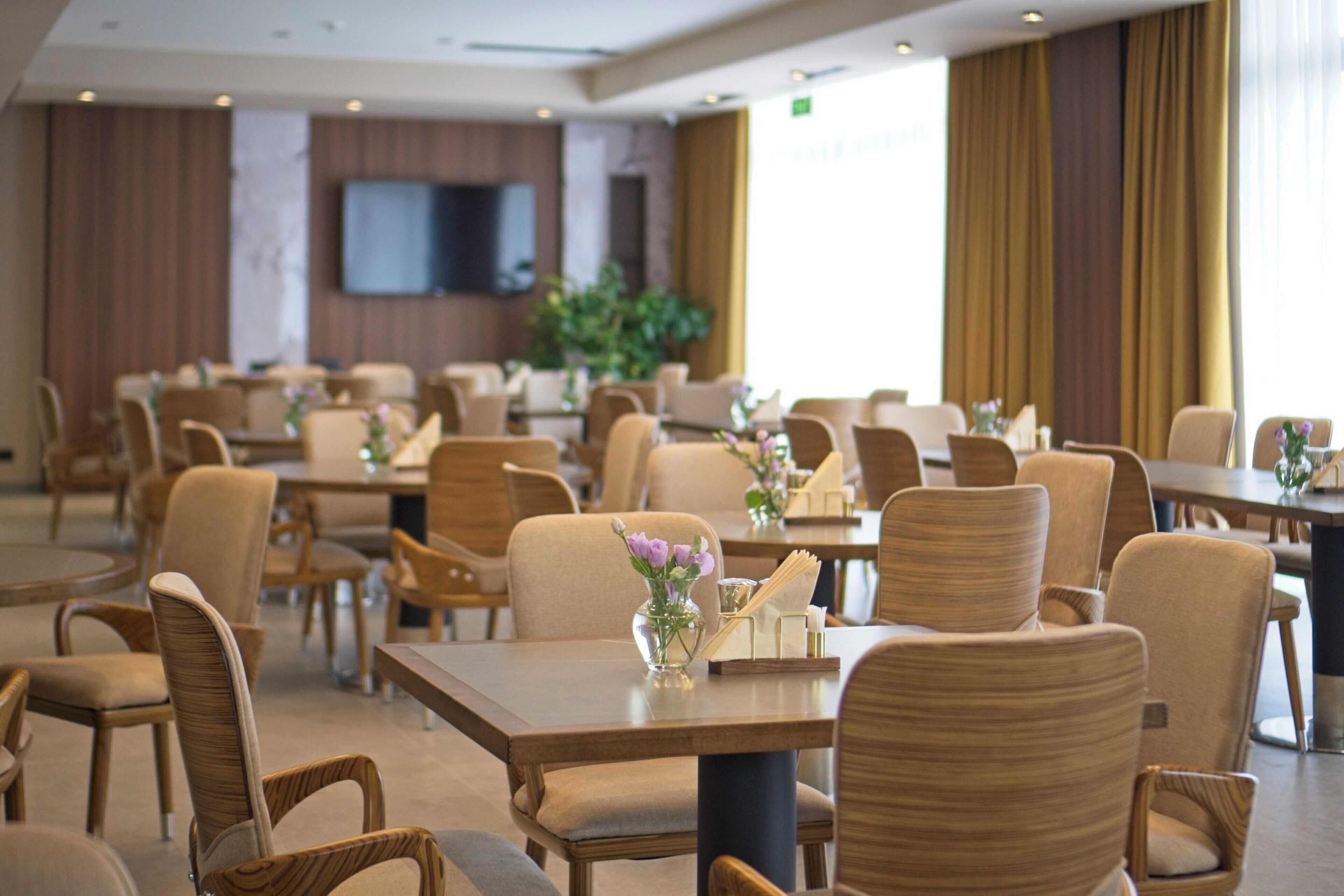 A spacious hotel dining area with multiple tables and chairs, a flat-screen TV mounted on the wall, and large windows with curtains.