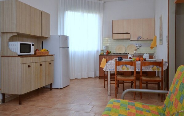 Cocina compacta con microondas, refrigerador y armarios de madera. Cuenta con una mesa de comedor con sillas y un cojín colorido.