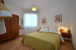 Un dormitorio con cama, armario, mesita de noche con lámpara y ventana con cortinas.