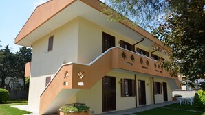 Un edificio de dos pisos con fachada color crema, una escalera que conduce a la entrada y un pequeño jardín con un banco.