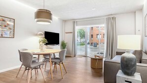 A modern hotel room with a dining area, a flat-screen TV, a sofa, a large vase, and a view of the street. - City Central Apartment w Modern Nordic Interiors (Aarhus)