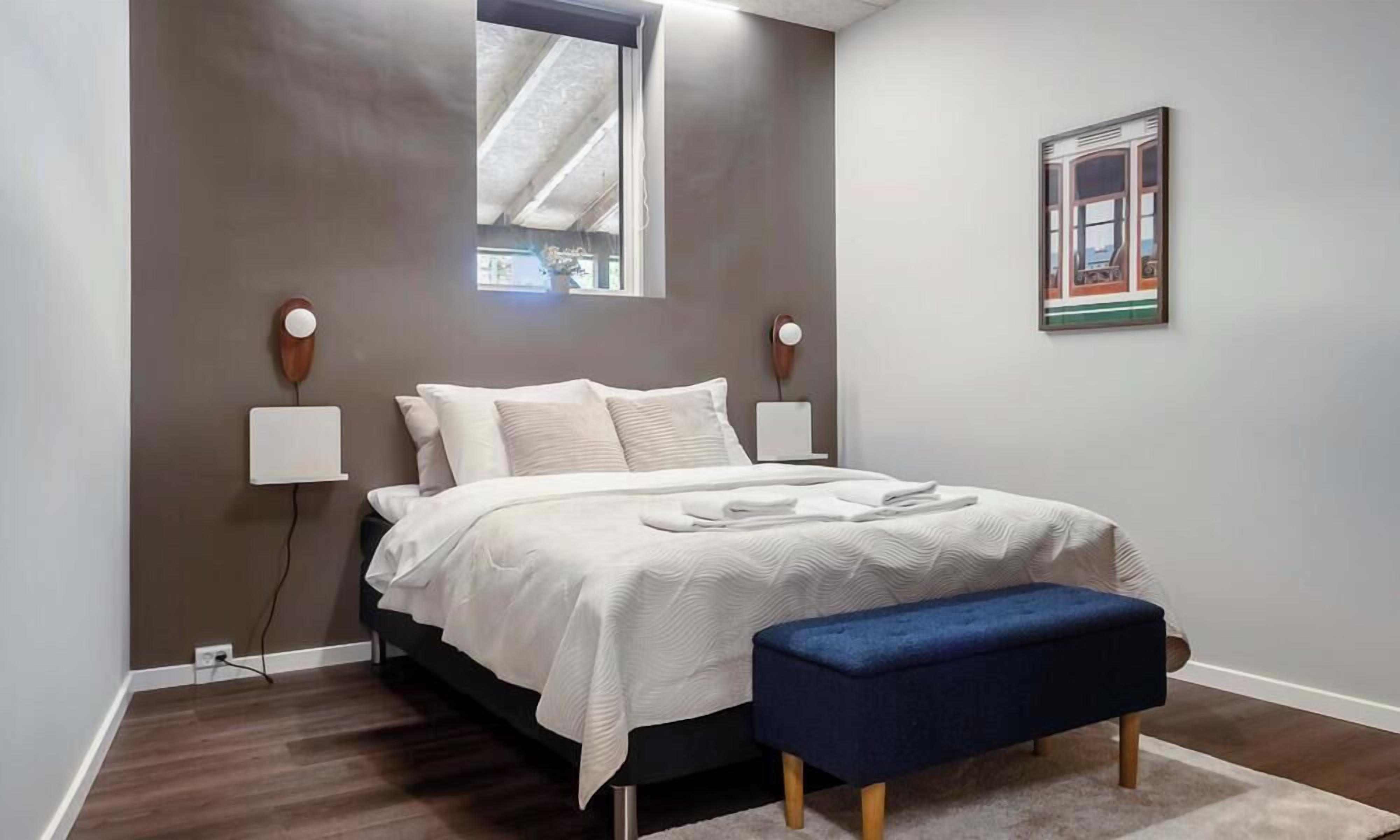 Een slaapkamer met een bed, nachtlampjes, een raam, een bankje en een lijst met een afbeelding aan de muur.