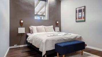 Een slaapkamer met een bed, nachtlampjes, een raam, een bankje en een lijst met een afbeelding aan de muur.