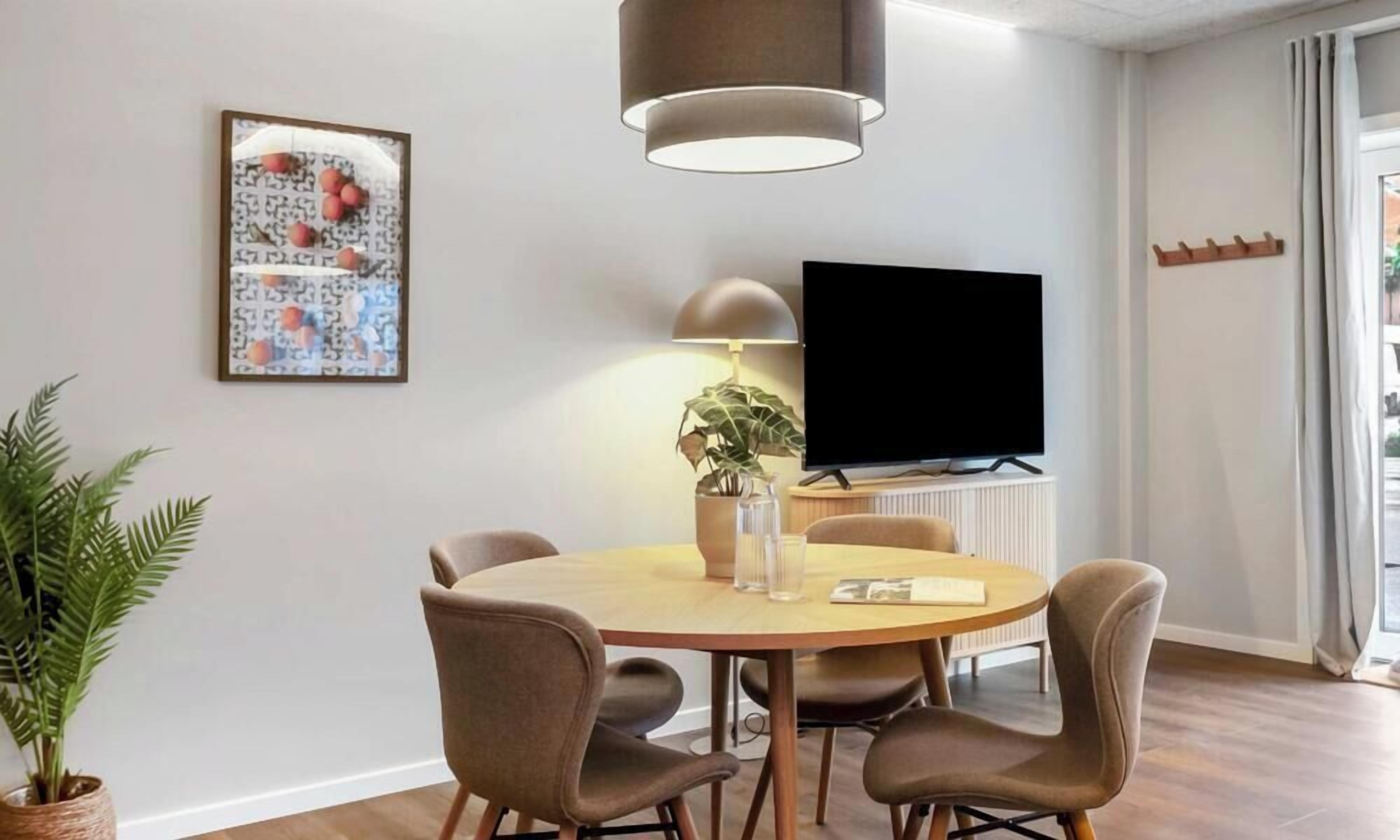 Een moderne eethoek met een ronde tafel, vier stoelen, een potplant, een flatscreen-televisie en een hanglamp.