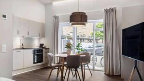 Een moderne keuken met witte kastjes, een eethoek met een ronde tafel en stoelen, een groot raam met gordijnen en een flatscreen-tv.