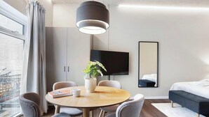 Een moderne hotelkamer met een ronde eettafel, grijze bekleden stoelen, een potplant, een flatscreen televisie, een grote spiegel en een bed met een donkere hoofdeinde.