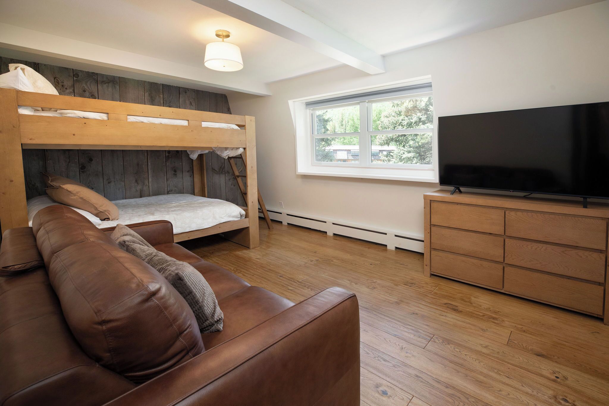 Quarto com cama beliche, sofá de couro marrom, televisão e janela com vista para árvores.