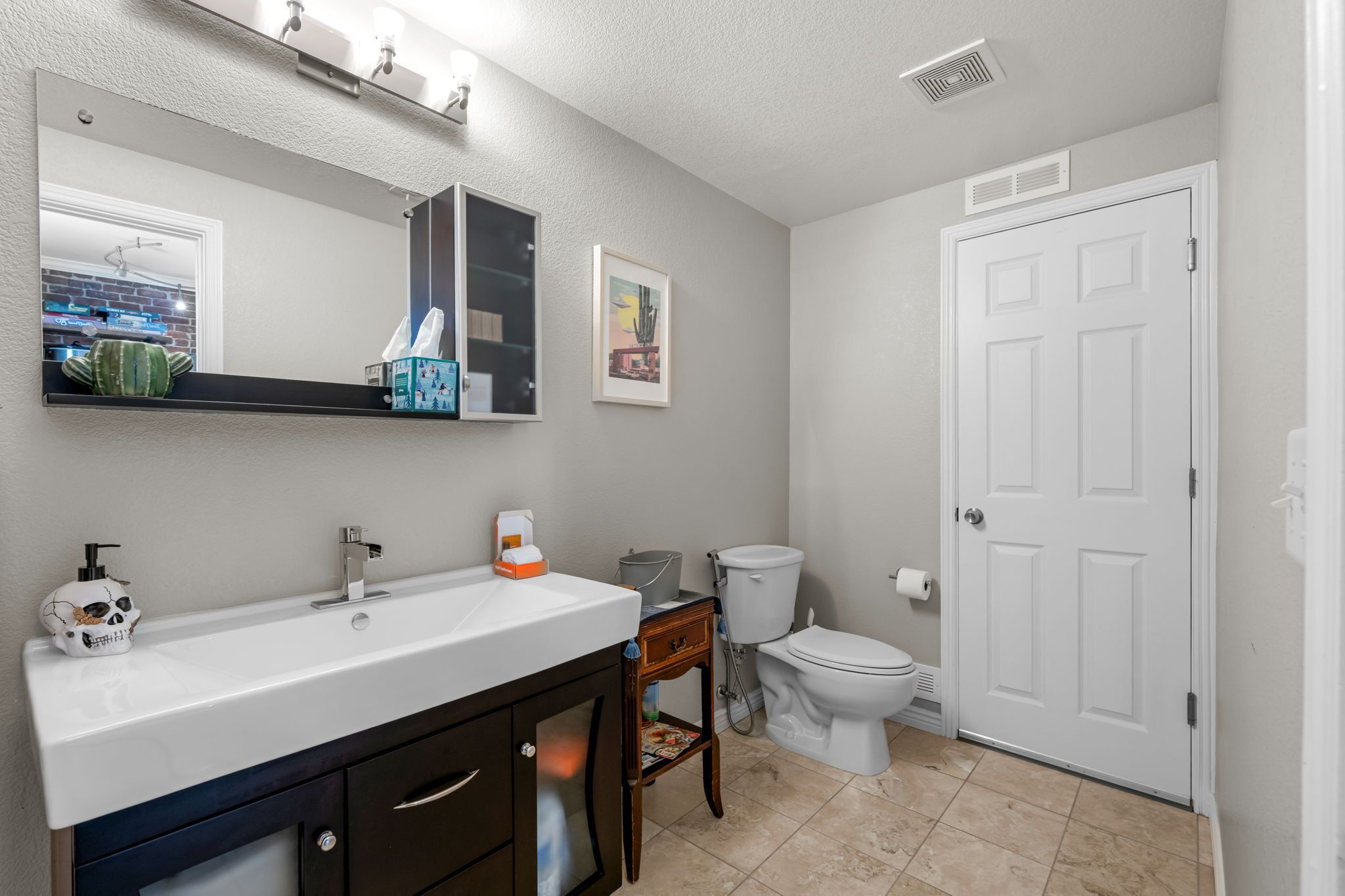 Un baño con lavamanos blanco, espejo e inodoro. Hay un mueble pequeño sobre el lavamanos y un cuadro en la pared.