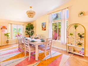 A dining room with a table set for six, surrounded by chairs, a chandelier, and a colorful rug. - Desert Oasis: 4BR Boho Oasis w/ Pool, Spa, Game Room & Sleeps 12 Comfortably (Indio)