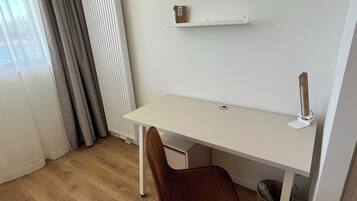 A small, neatly arranged room with a white desk, a brown chair, a white radiator, and a window with curtains.