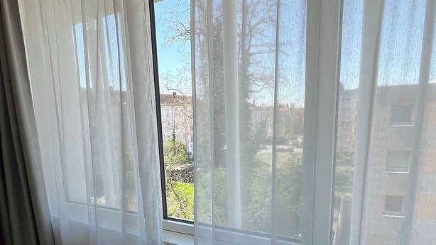 Una stanza con un'ampia finestra coperta da tende bianche trasparenti, pavimento in legno e vista su edifici e alberi.