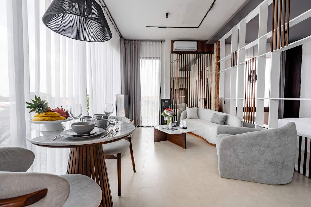 A modern living room with a round dining table, grey sofas, and a large window with sheer curtains.