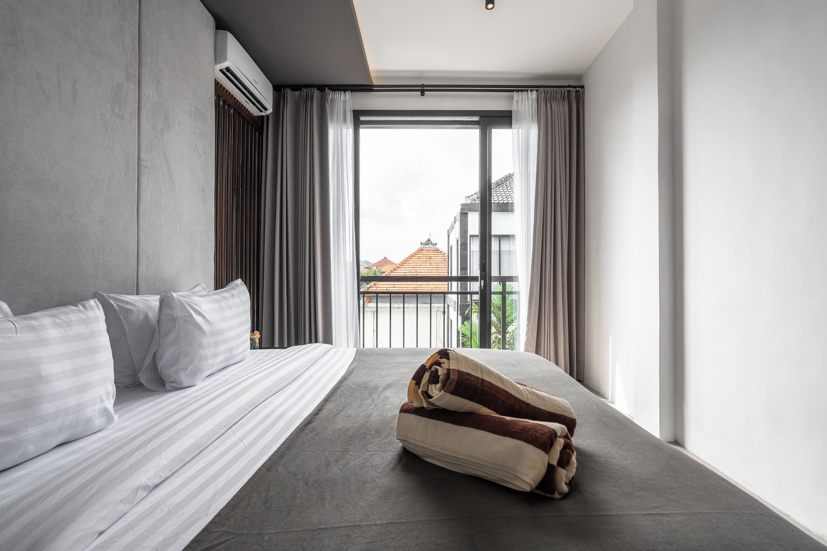 A modern hotel room with a large bed, a view of the city through a balcony, and an air conditioning unit on the wall.