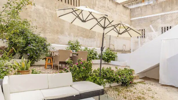 An outdoor seating area with a white sofa, a table, and chairs, surrounded by potted plants and a large umbrella.