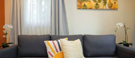 A grey sofa with two cushions, a white curtain, and a painting of plants on the wall.