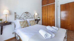 Un lit bien fait, avec du linge de lit blanc et des coussins décoratifs, une armoire en bois et deux lampes de chevet.