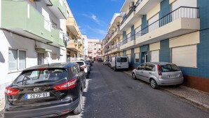 Une rue étroite en milieu urbain, bordée d’immeubles résidentiels à plusieurs étages, de voitures garées et d’un ciel bleu dégagé.