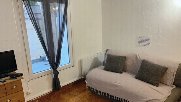 Ein Zimmer mit einer Couch, einem Fernseher auf einem Holzkasten und einem Fenster mit Vorhängen.