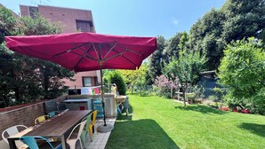 A patio area with a wooden table and chairs, a red umbrella, and a small garden with a lawn and potted plants. - Oasis beach & golf Apartment nestled in nature next to the beach (Venezia)