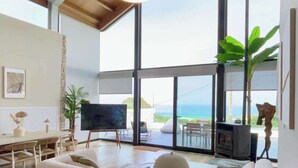 A spacious living room with a high ceiling, large windows offering a view of the ocean, a modern fireplace, and a comfortable seating area.
