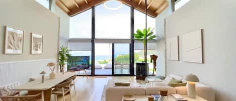 A spacious living room with a high ceiling, large windows offering a view of the sea, a dining area with a table and chairs, and a cozy seating area with a sofa and cushions.