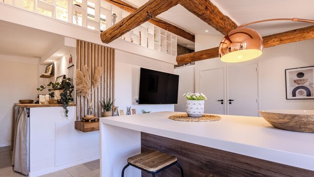 A modern kitchen with a central island, wooden beams, a mounted TV, and a pendant light.