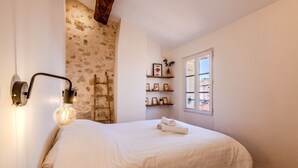 A bedroom with a large bed, a wall-mounted light fixture, a wooden ladder, and a window with a view.