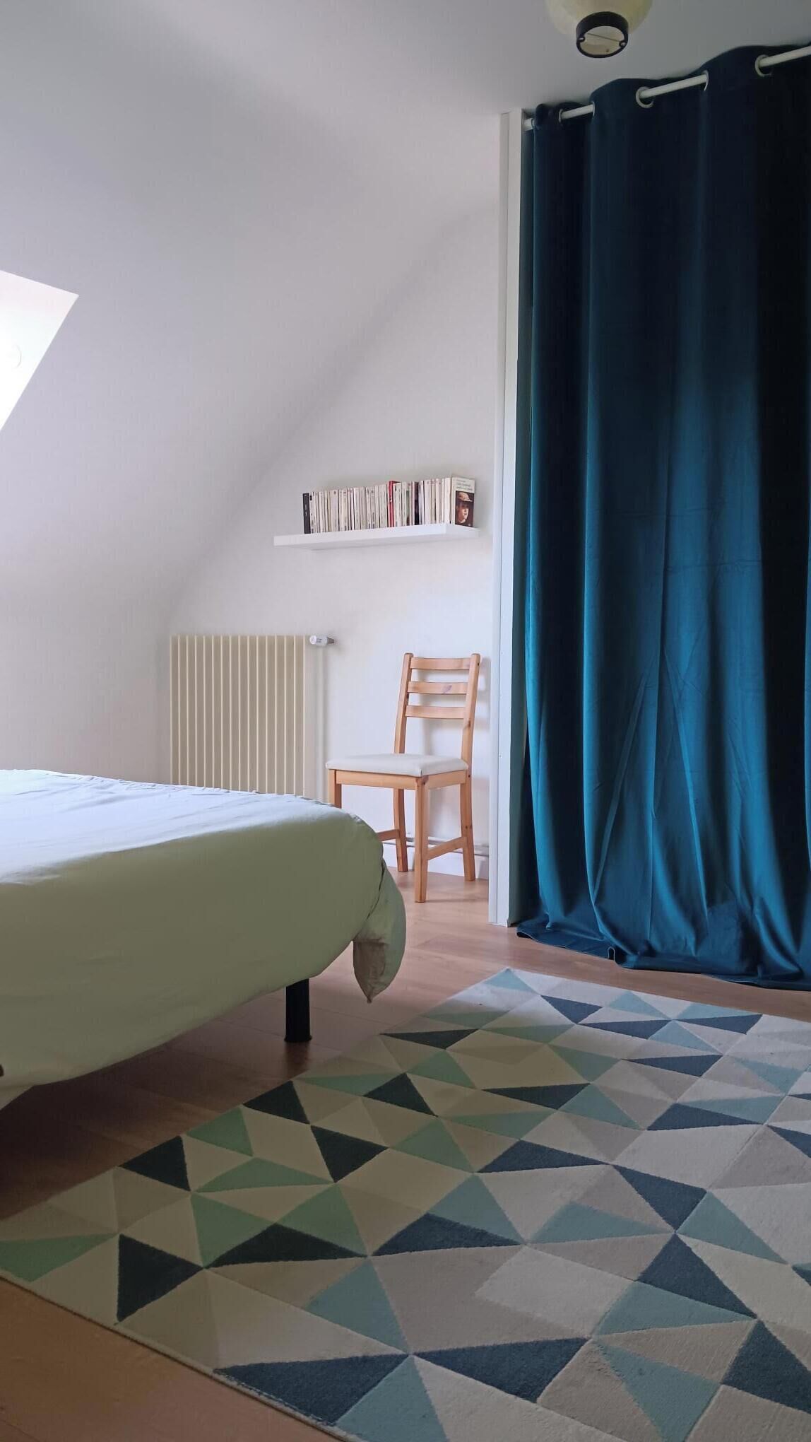 Ein Zimmer mit einem Bett, einem Sessel, einem Bücherregal und einem Fenster mit Vorhängen.