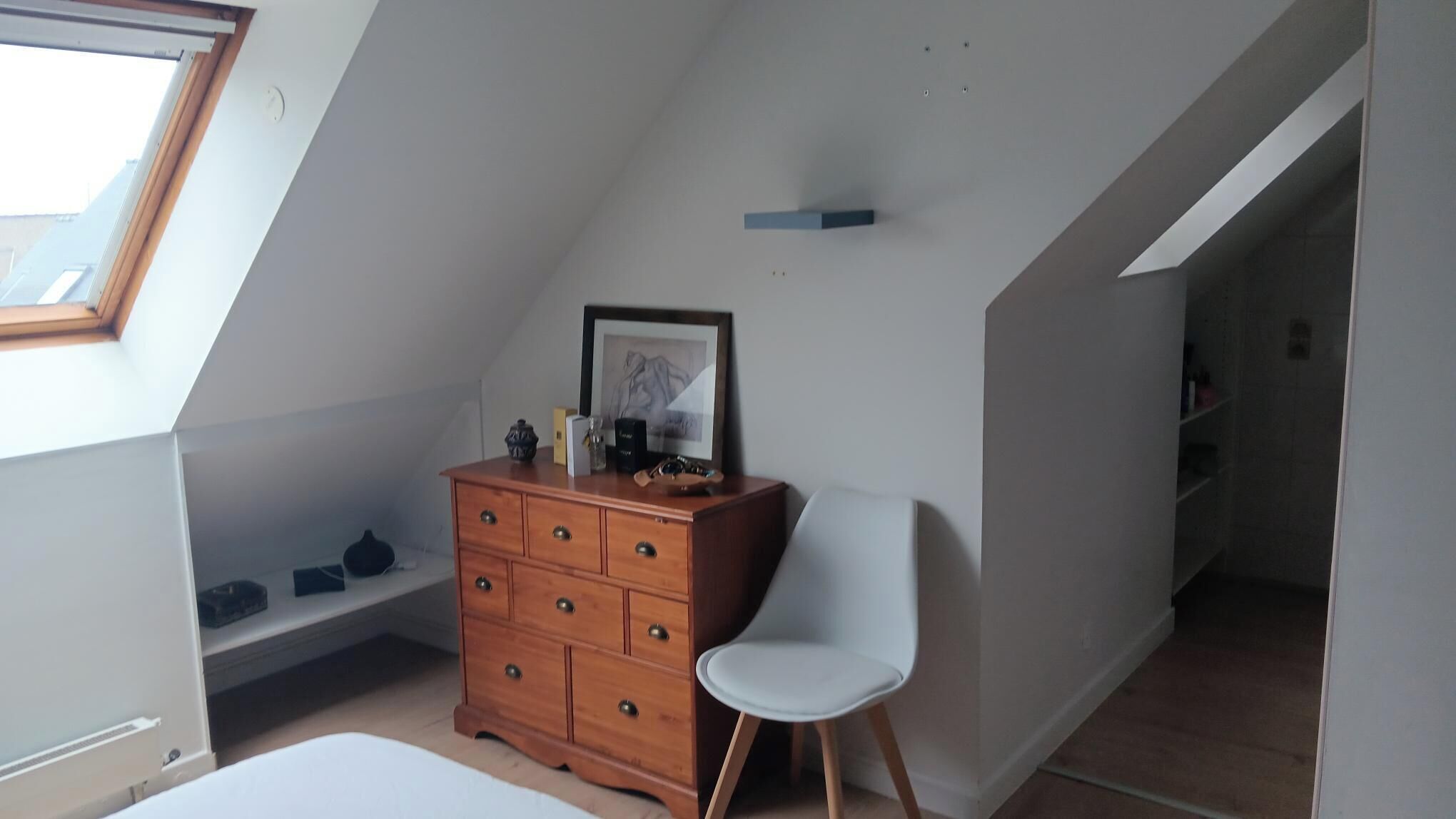 A small, sloped room with a wooden dresser, a white chair, and a framed picture on the wall.
