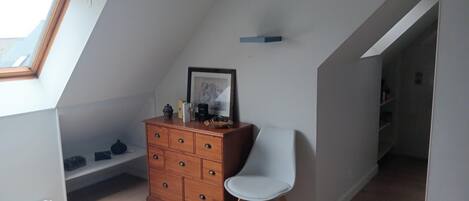 A small, sloped room with a wooden dresser, a white chair, and a framed picture on the wall.
