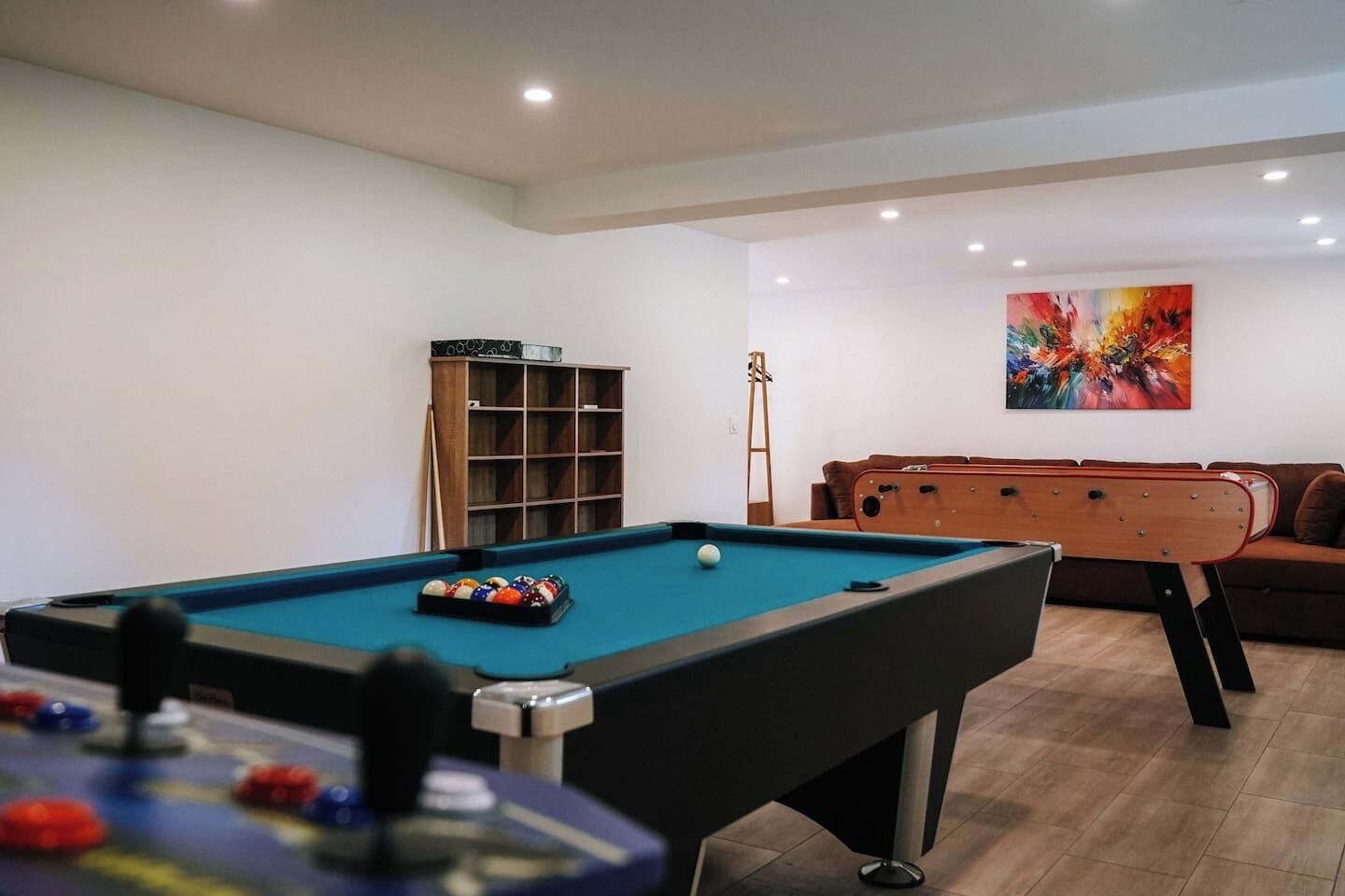 A pool table with balls, a cue stick, and a billiards table in a room with a brown sofa and a colorful painting on the wall.