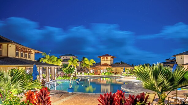 Área de piscina en un resort de noche, con vista despejada de la piscina, follaje circundante y edificios cercanos.