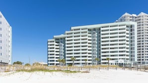 Un hotel de varios pisos con balcones, una playa arenosa en primer plano y un cielo azul despejado.