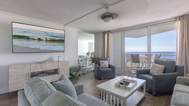 Un salón moderno con un televisor de pantalla plana grande, sofás grises, una mesa de centro blanca y un balcón con vistas al mar.