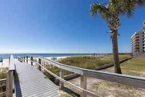 A wooden boardwalk leading to a beach with a palm tree and a building in the background. - Perdido Sun 516- Stunning Large Gulf Front Luxury (Pensacola)